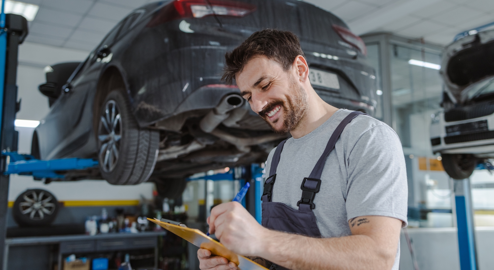 A mechanic smiles while taking notes on a clipboard in an auto repair shop, with a car raised on a lift in the background.