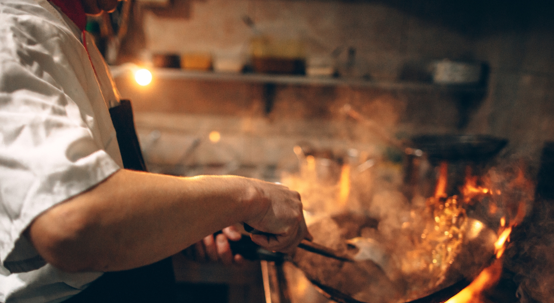 A chef cooking in a professional kitchen with a wok over an open flame.