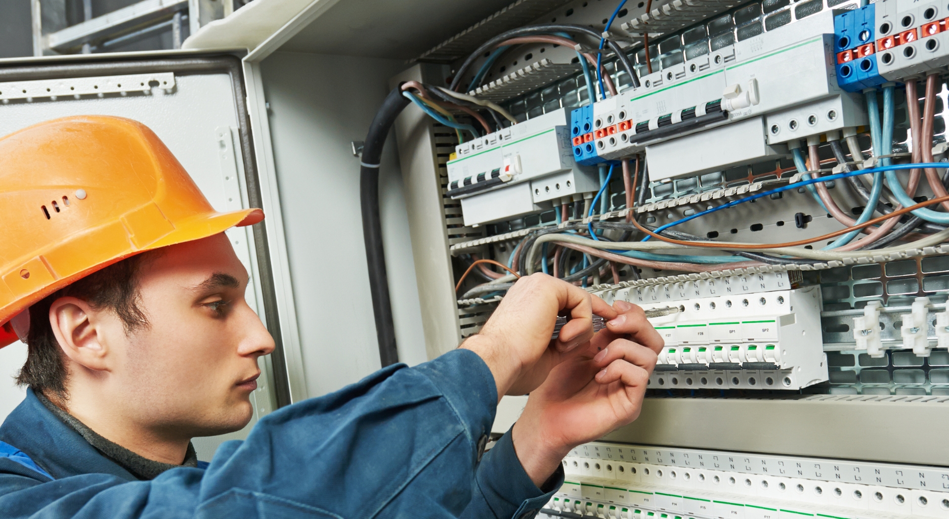 Electrician in a hard hat working on an industrial electrical panel, focusing on wiring and circuit breakers.