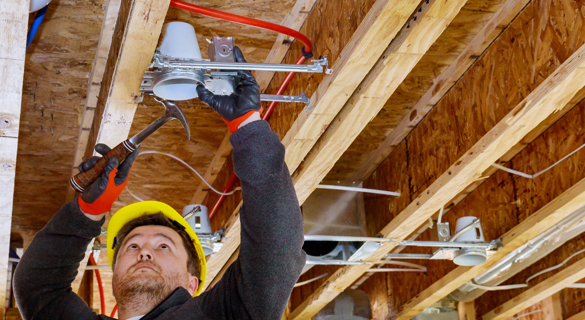 A construction worker wearing a yellow hard hat and gloves installs electrical fixtures on exposed wooden beams in an industrial setting.