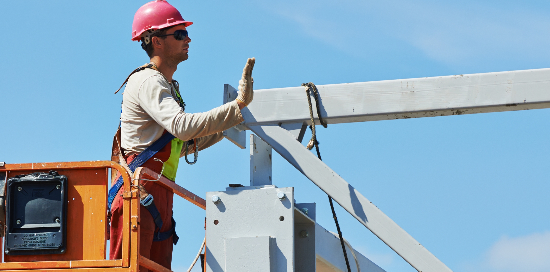 Construction worker in a lift wearing a pink hard hat and safety gear, working on a metal structure, exemplifying a millwright's tasks under a clear blue sky.