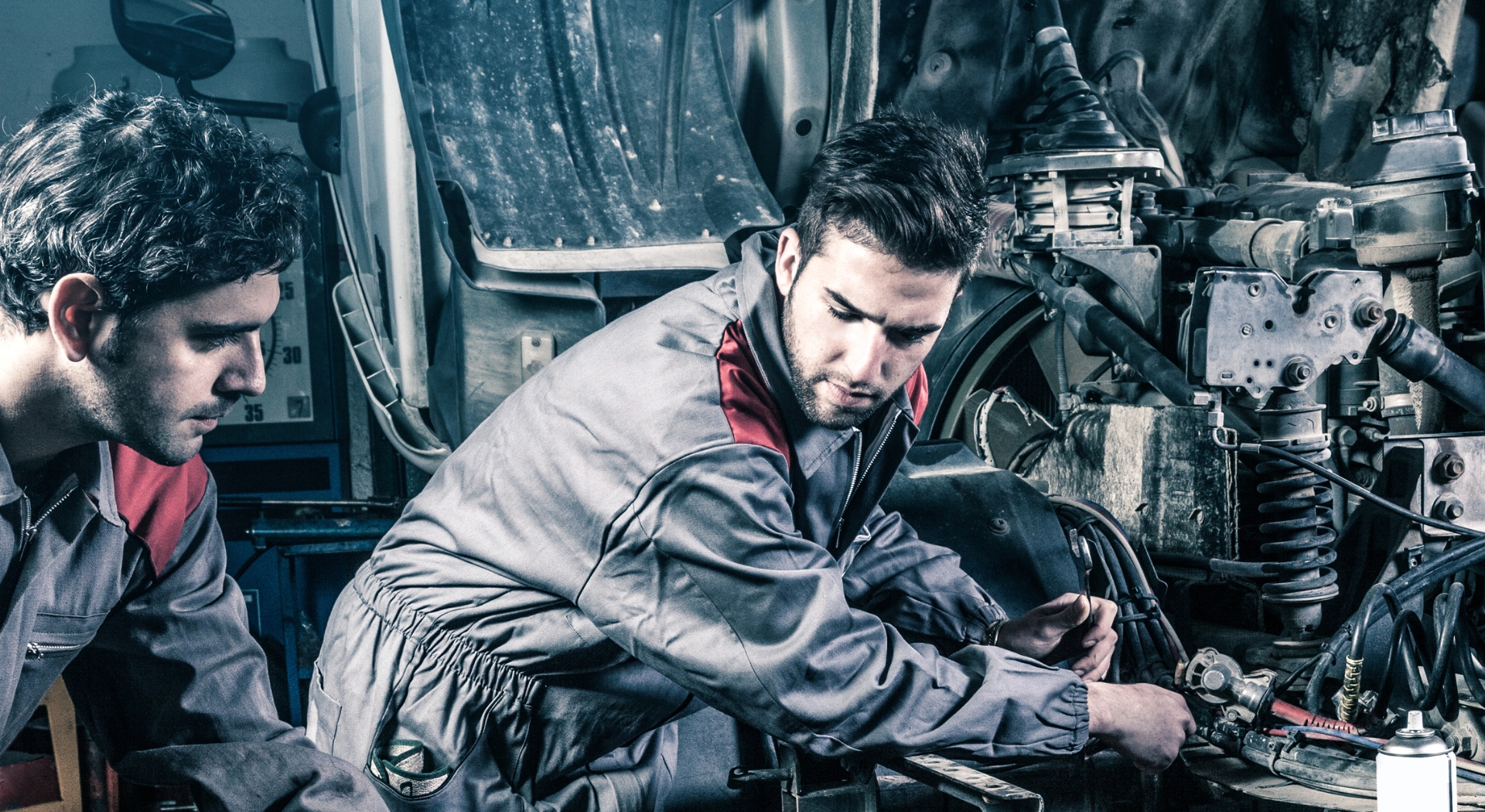 Two mechanics working on a truck engine, wearing gray overalls, in a workshop setting.