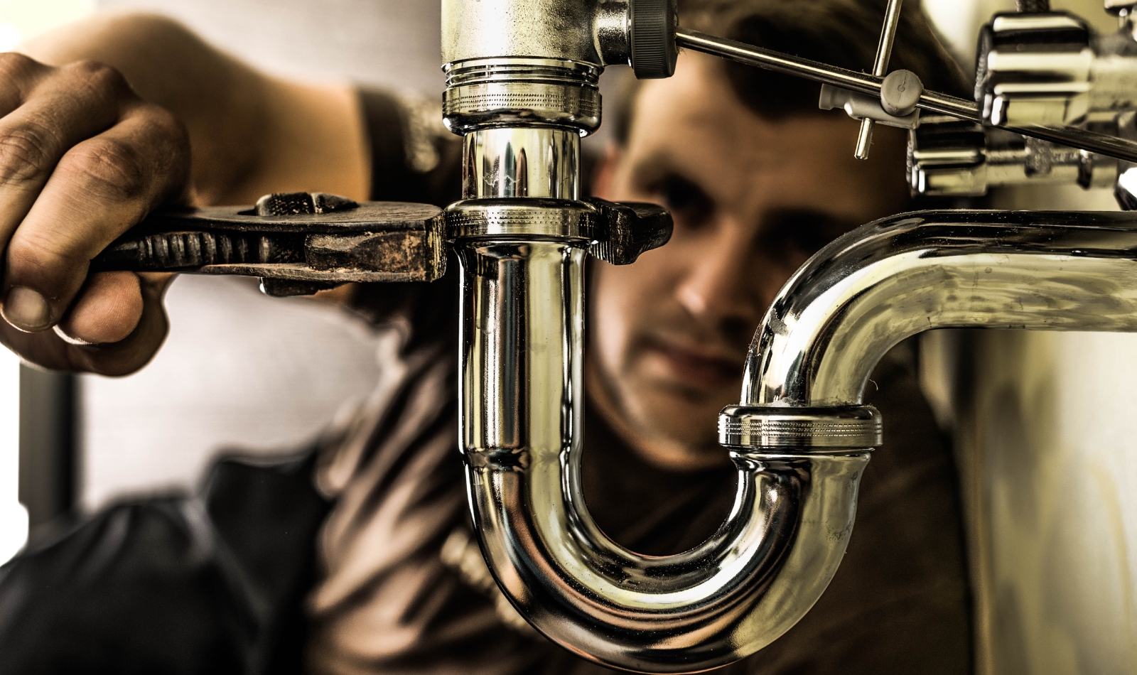 A plumber using a wrench to adjust a chrome pipe under a sink.