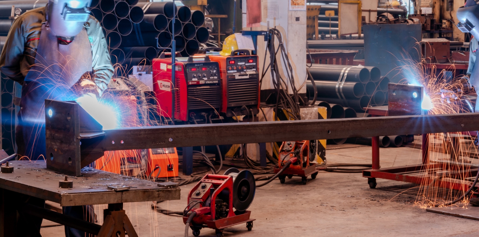 Welder working in an industrial workshop, creating sparks during the welding process, surrounded by equipment and metal pipes.